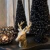 decoratief herten hoofdje van 12,5 cm hoog. gemaakt van keramiek. De kleur is antiek goud.Op de foto gedecoreerd op een zwart metalen plateau in een organische vorm. Erbij staat een zwarte metalen waxinehouder met een opengewerkte structuur. Daarnaast staan twee zwarte kerstboompjes van verschillende hoogtes. Deze boompjes hebben een mooie sparkle.